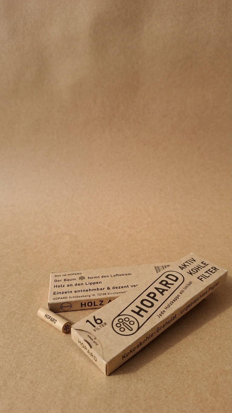 zwei Flache Faltschachtelen aus Kraftpapier mit 16 Filtern, ein Filter wird einzeln entnommen und liegt seitlich auf dem Kraftpapier daneben